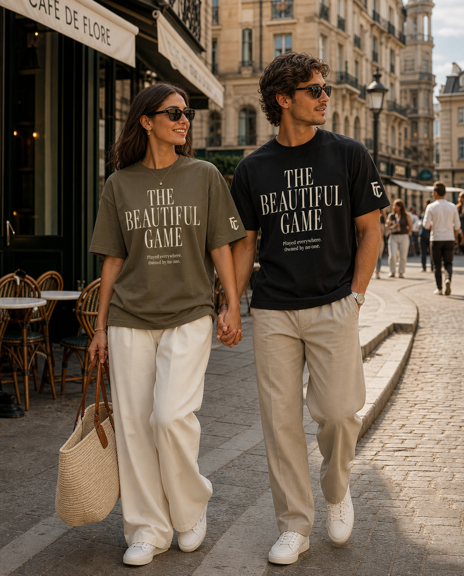 A couple walking hand-in-hand down a cobblestone street in Paris, wearing matching graphic t-shirts that read 'THE BEAUTIFUL GAME' in white text representing World Cup soccer. The woman wears an olive green shirt with white pants, and the man wears a black shirt with beige pants.