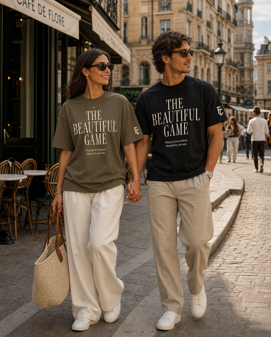 A couple walking hand-in-hand down a cobblestone street in Paris, wearing matching graphic t-shirts that read 'THE BEAUTIFUL GAME' in white text representing World Cup soccer. The woman wears an olive green shirt with white pants, and the man wears a black shirt with beige pants.